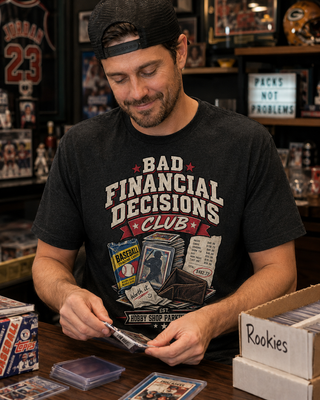 A man wearing a black graphic t-shirt with 'Bad Financial Decisions Club' printed on the front, sitting at a wooden table surrounded by trading cards and card sleeves in a collector's room.