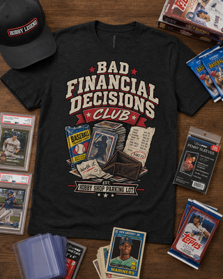 A black graphic t-shirt featuring a retro-style baseball print that reads 'BAD FINANCIAL DECISIONS CLUB' with illustrations of baseball cards, a wallet, and a receipt. The shirt is styled in a flat-lay arrangement on a wooden table alongside a black 'Robbery Landing' cap, trading cards, and baseball card packs.