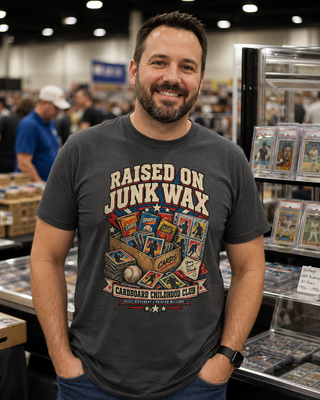 A smiling man wearing a dark gray graphic t-shirt with 'RAISED ON JUNK WAX' and a cardboard children's club design, standing at what appears to be a card convention or retail display.