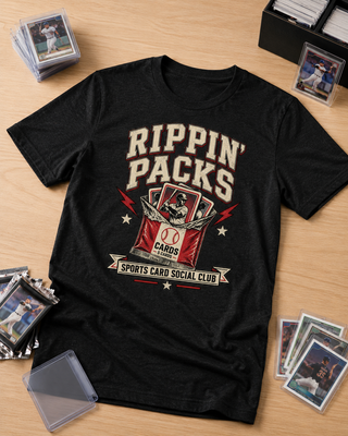 A black graphic t-shirt laid flat on a light wood surface, surrounded by trading cards and card sleeves. The shirt features a distressed cream and red graphic that reads 'Rippin' Packs' and 'Sports Card Social Club'.