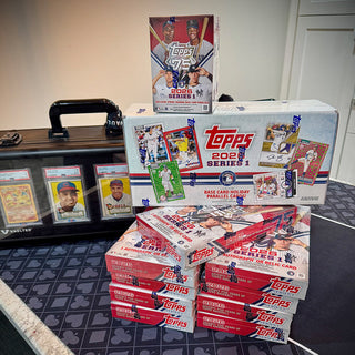 Stack of Topps trading card boxes on a table with a display case and cards in the background.