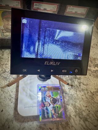 A handheld ELIKLIV card reader microscope displaying a blue-toned scan of a collectible trading card, with the card visible below on a white stand over a granite countertop.